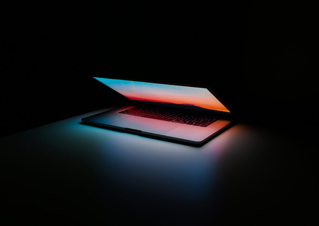 A slightly open laptop on a desk in an unlit room. A blue and orange glow emits from the screen's background of a sunset.