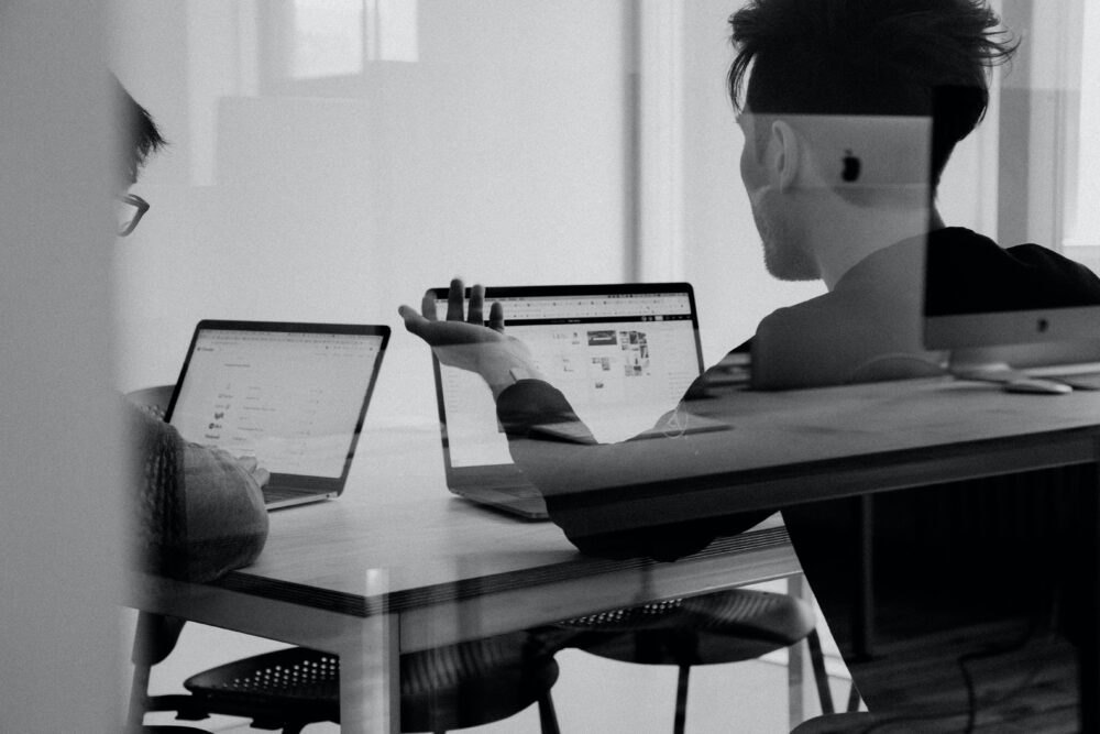 In black & white, two people are seen through glass sitting at a table working together with 2 laptops.