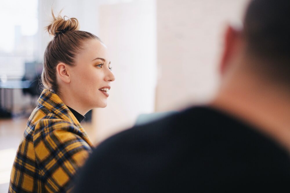 A woman in a yellow plaid shirt with a bun speaks to others, with a man in the foreground listening.