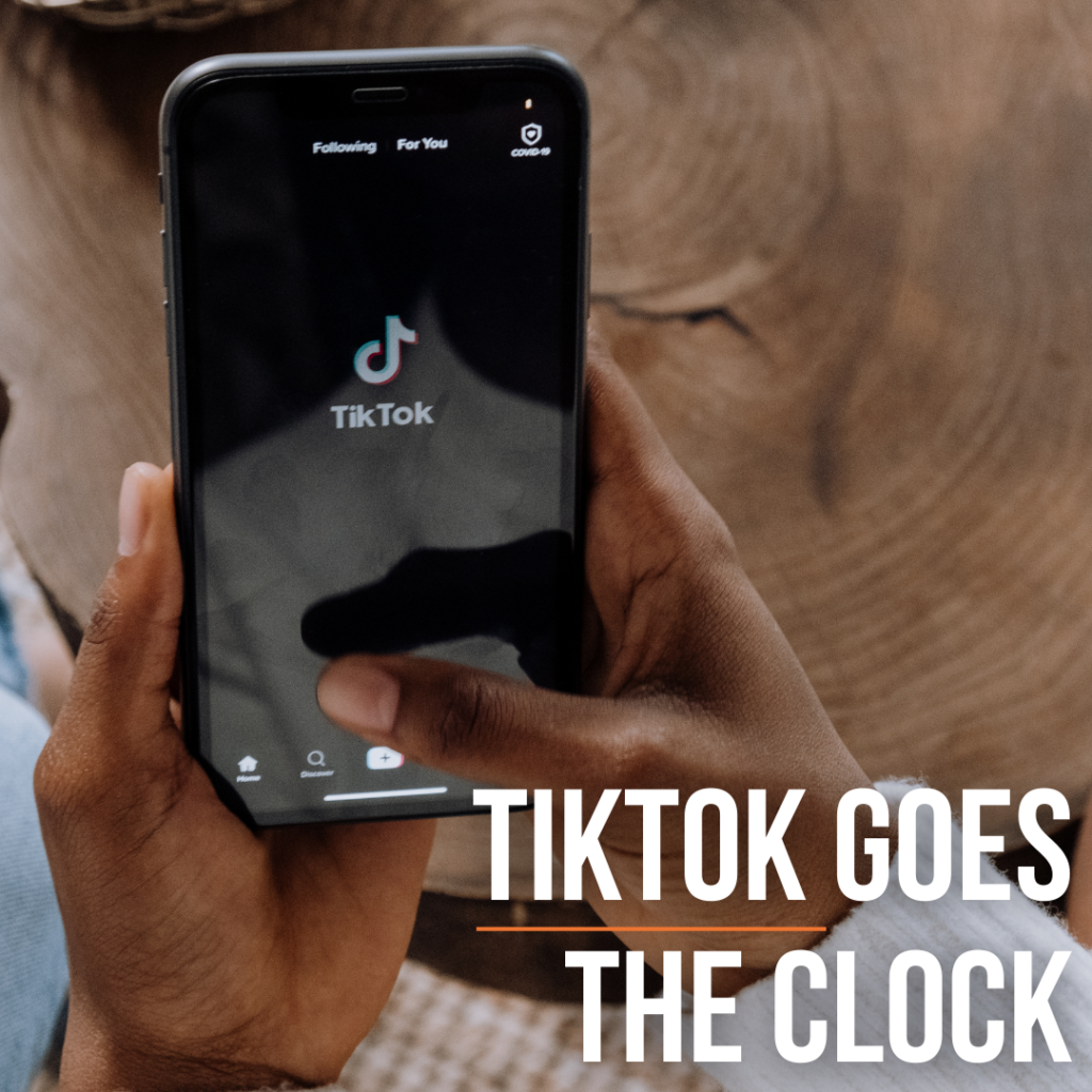 A Black pair of hands hold a phone displaying the TikTok logo above a natural wood edge table. Text: "TikTok goes the clock."