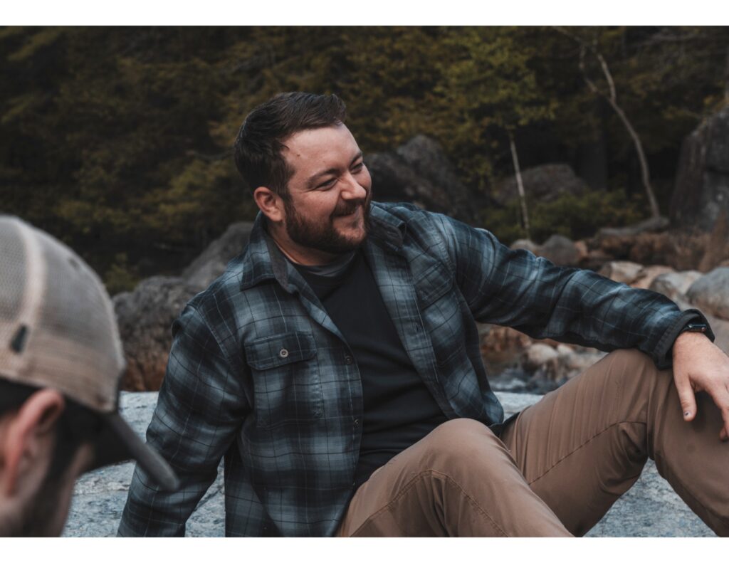 Charles McDonald sits atop a boulder in a lush forest. He's wearing a faded blue plaid shirt, brown pants, a watch, and a winning smile peeks from beneath his beard.